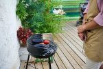 Schwarzer Griller auf einer Terrasse mit Holzboden. Davor steht ein Mann in Grillschürze. © vecteezy.com