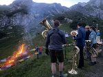 Personen spielen auf Blechblasinstrumenten vor einer Bergkulisse. Im Hintergrund brennt ein kleines Feuer.