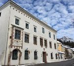 Historisches Gebäude mit weißer Fassade und dunklen Fensterrahmen. Im Hintergrund Berge und blauer Himmel.