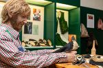Person arbeitet konzentriert an einem Lederstück an einem Holztisch. Im Hintergrund sind Ausstellungsstücke in Vitrinen zu sehen.
