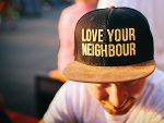 Eine Person trägt eine schwarze Kappe. Auf der Kappe steht in gelber Schrift: „LOVE YOUR NEIGHBOUR“. Das Gesicht der Person ist nicht zu erkennen. 
